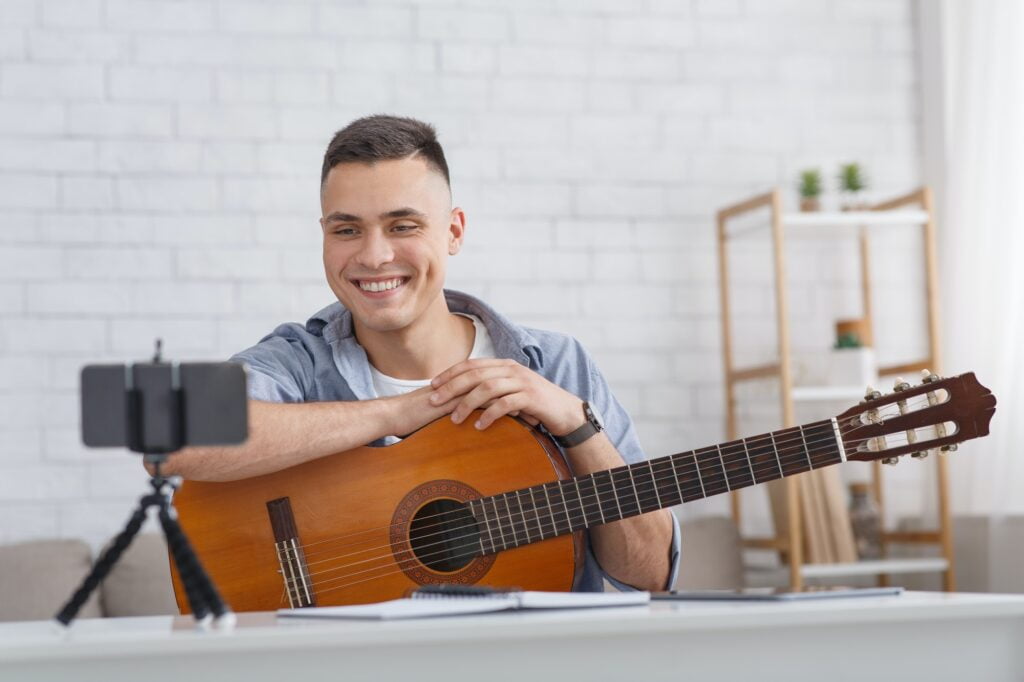 Online music lesson. Smiling guy with guitar looks at smartphone camera