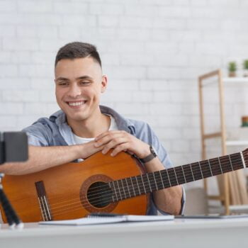 Online music lesson. Smiling guy with guitar looks at smartphone camera